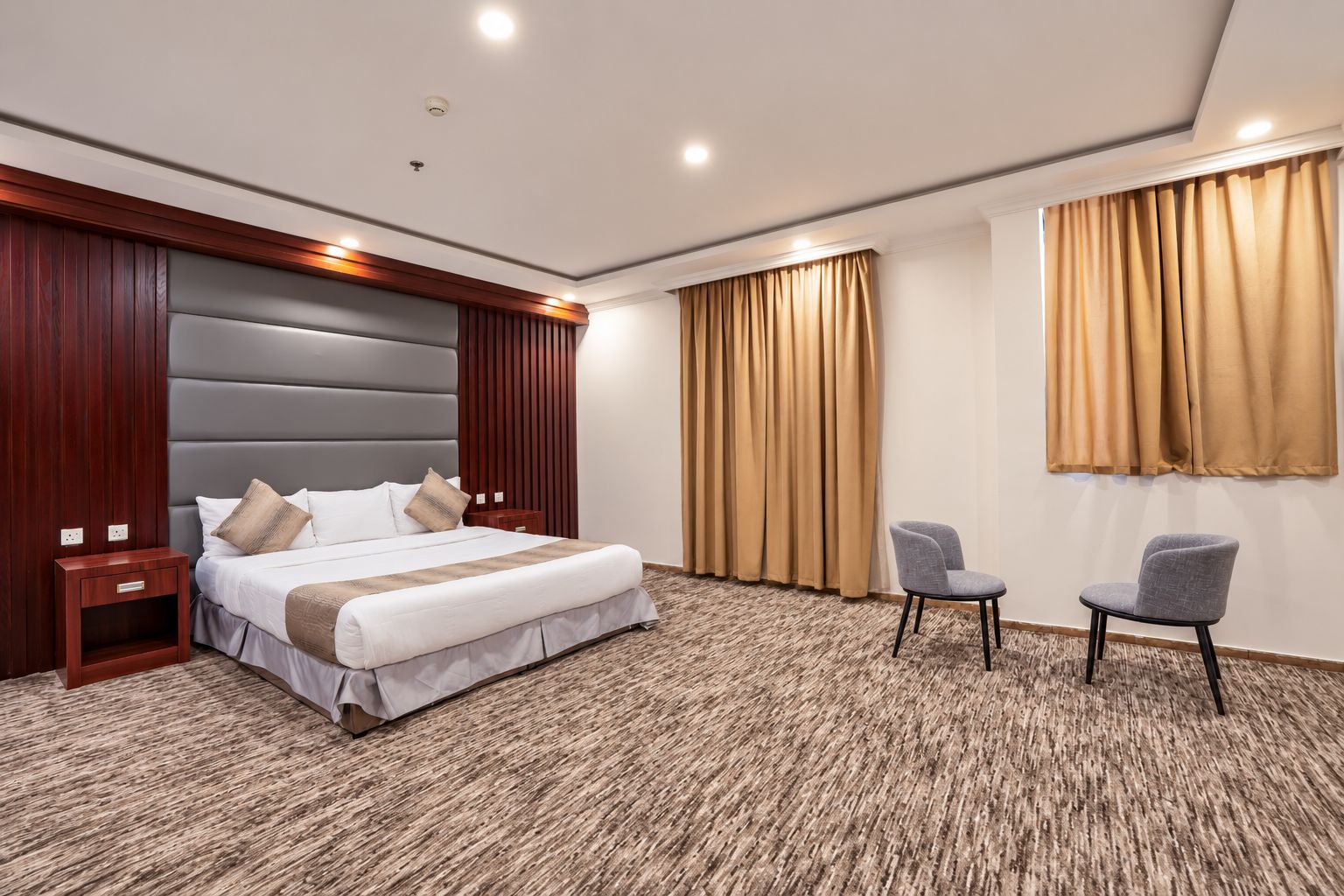 A hotel room with a large bed, a wooden headboard, a small desk, two chairs, and a large window with curtains.