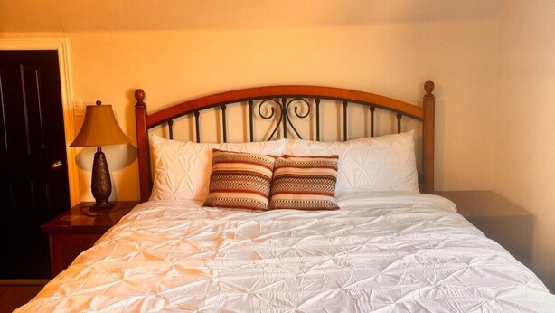 Un lit bien fait, avec une couette blanche et deux coussins à motifs, une tête de lit en bois, une lampe de chevet et une porte sombre.