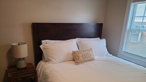 A neatly made bed with white linens and a patterned pillow, a wooden nightstand with a lamp, and a window with blinds.