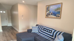 A living room with a dark blue sectional sofa, a round wooden coffee table, a patterned rug, and a painting of a coastal scene on the wall.