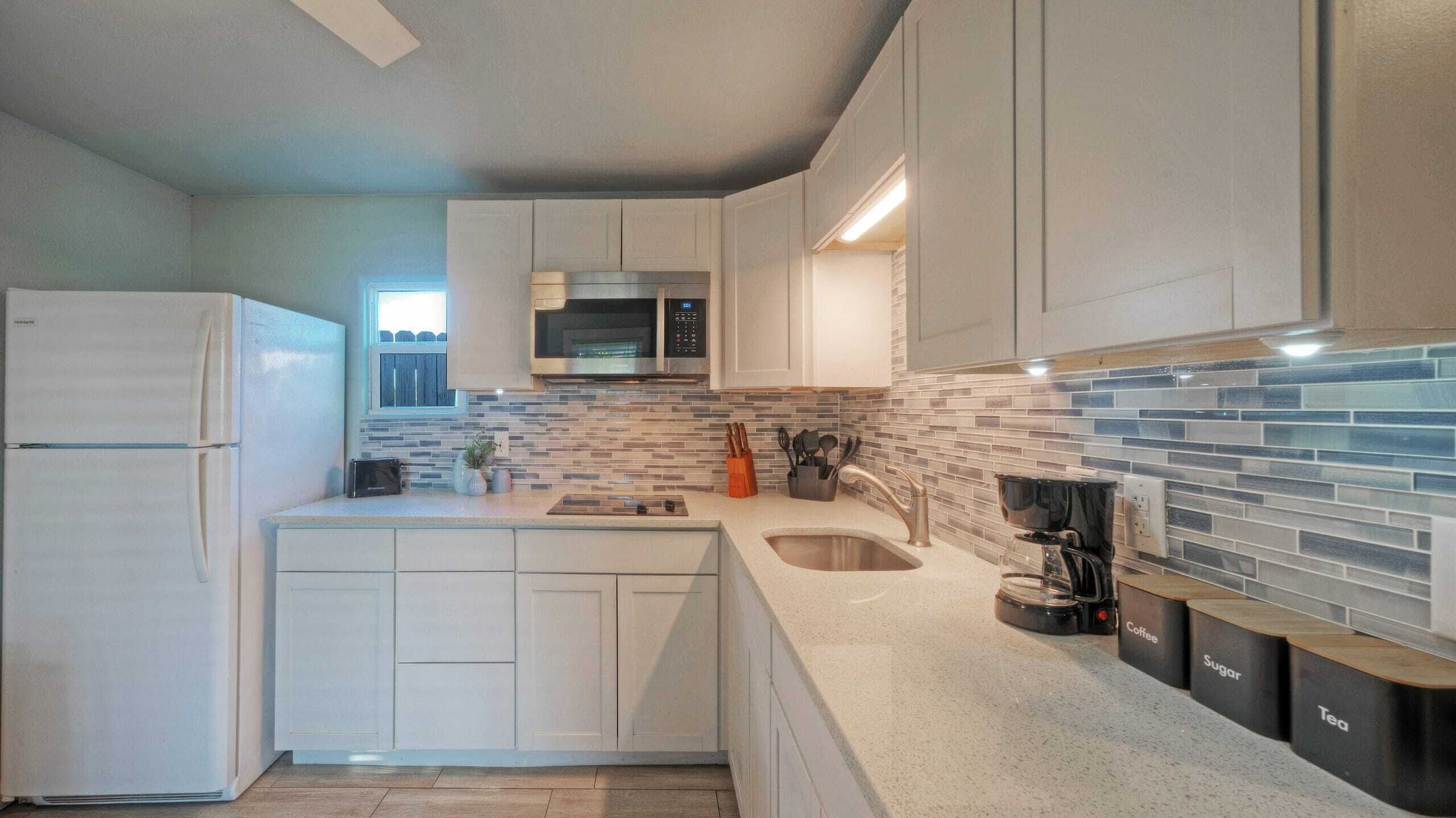 Una cocina moderna con gabinetes blancos, un microondas de acero inoxidable, una cafetera y un salpicadero de azulejos.