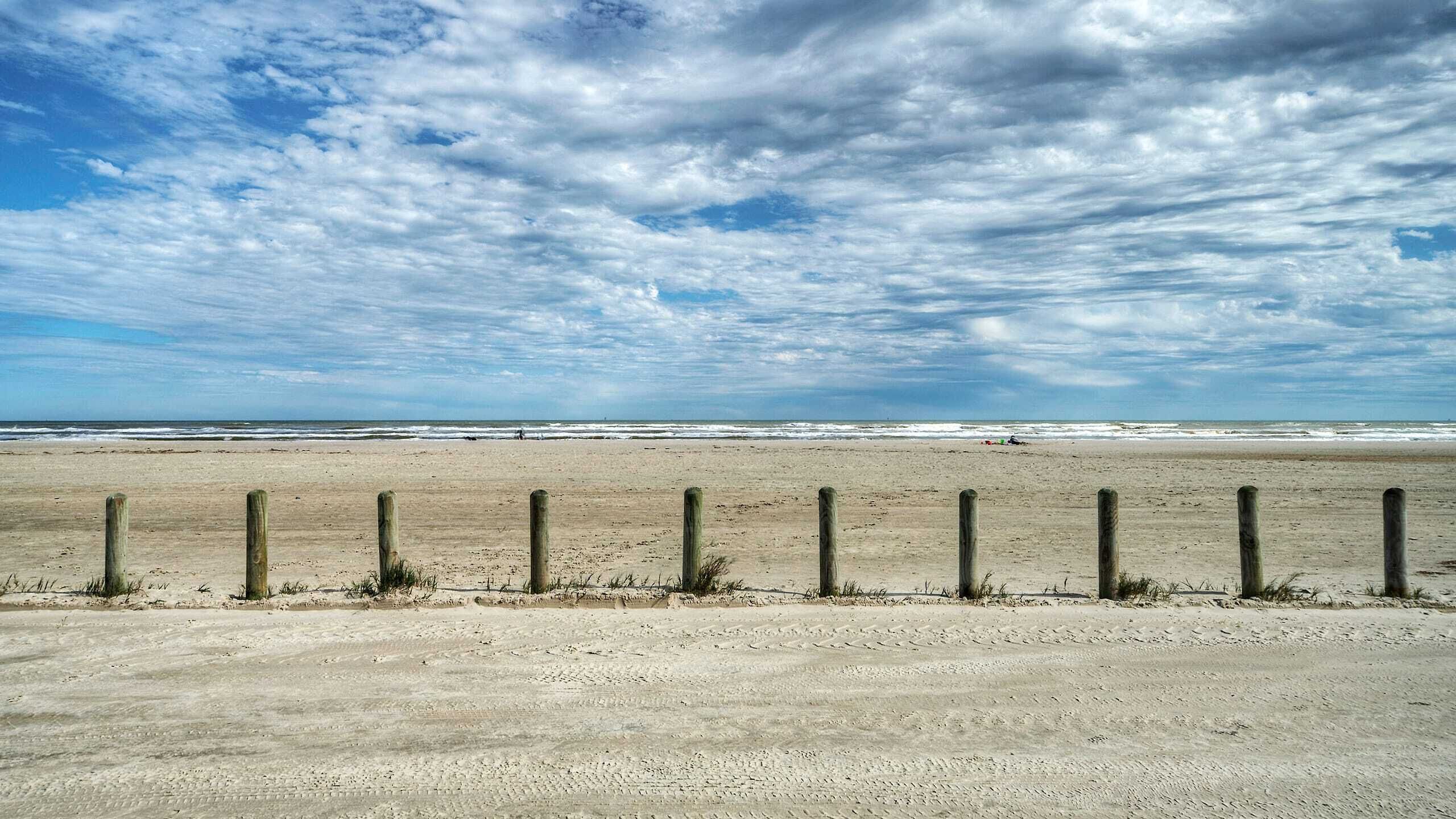 Una playa arenosa con una valla de madera en primer plano y el océano al fondo.