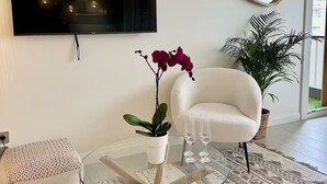 Un salón moderno con un televisor de pantalla plana, un espejo redondo, un sillón blanco, una mesa de centro de vidrio con una planta en maceta y una alfombra estampada.