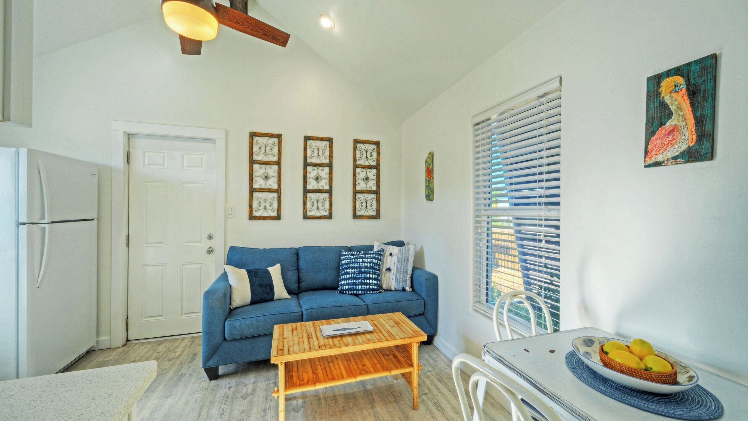 A compact living space with a sofa, a wooden coffee table, a refrigerator, and a dining table with a fruit bowl.