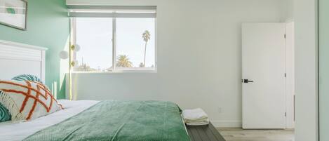 A bedroom with a bed, a bench, a window with a view of palm trees, and a white door.