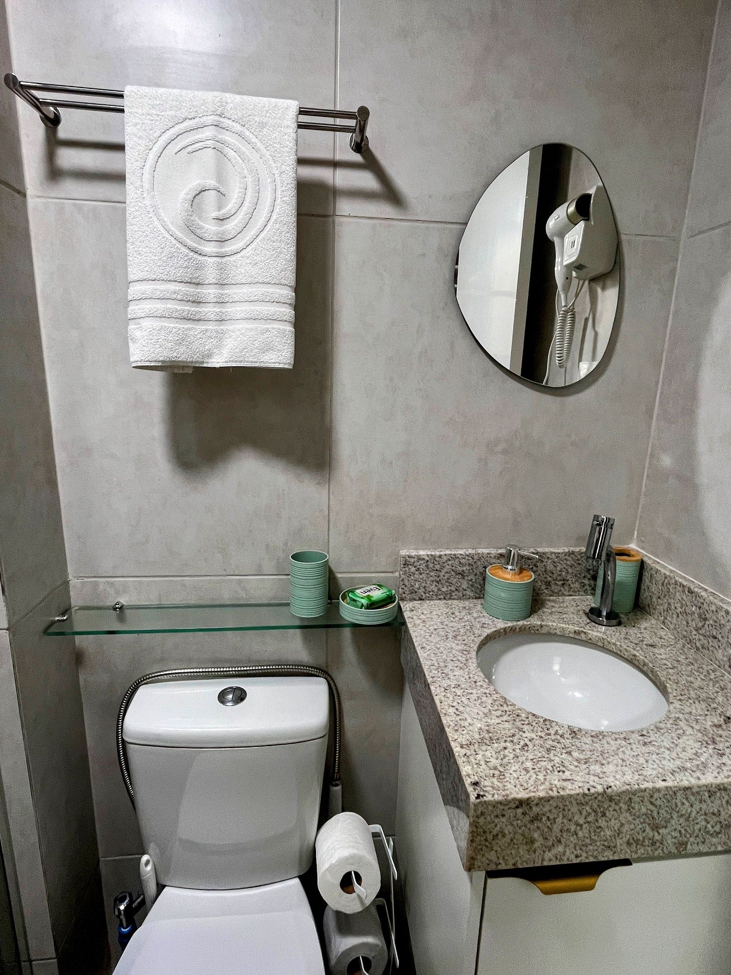 Baño con inodoro, lavabo, espejo, toallero y estante de vidrio con artículos de aseo.