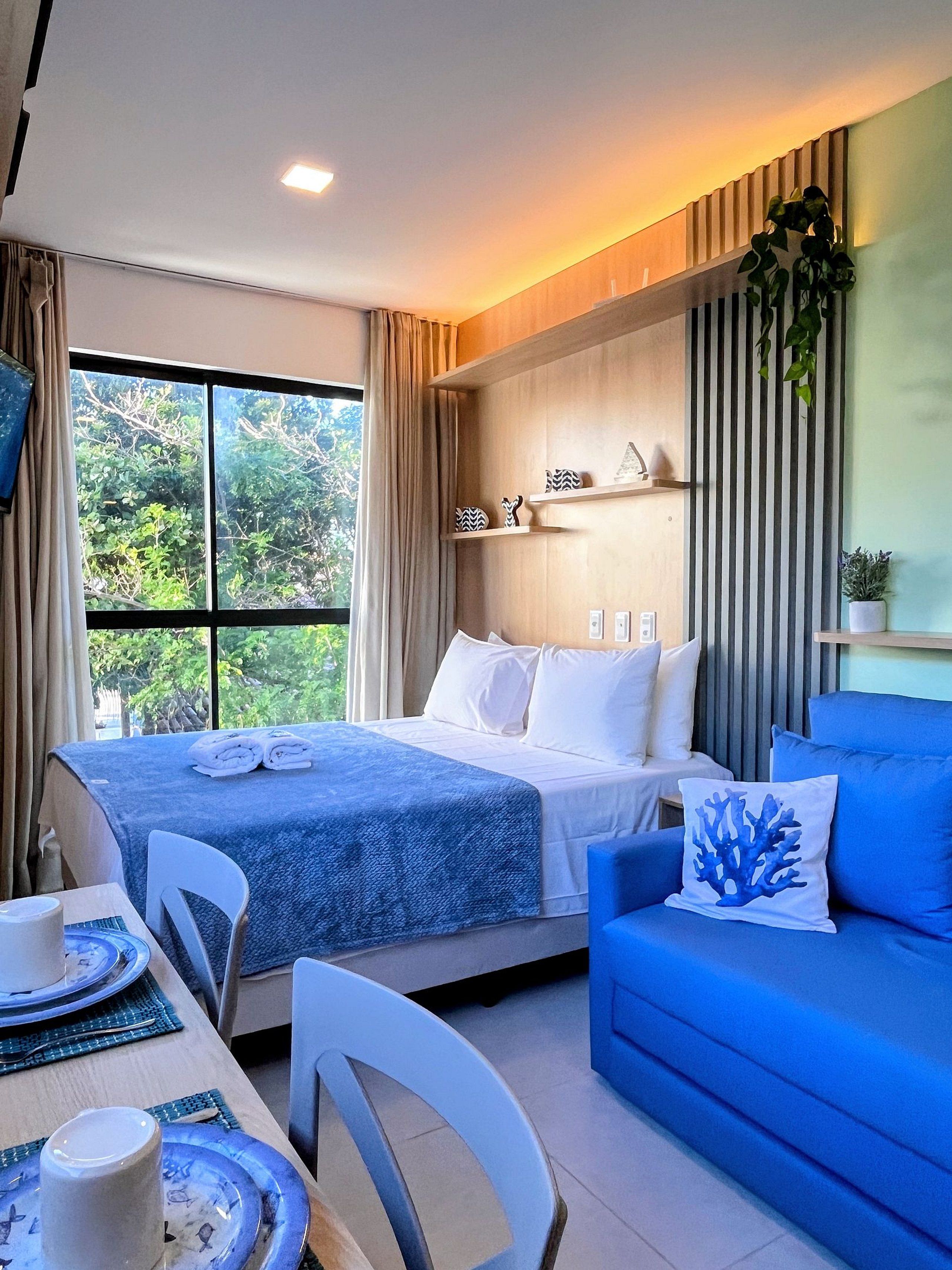 Quarto de hotel com cama, sofá azul, mesa de jantar com pratos e xícara, e janela com cortinas.
