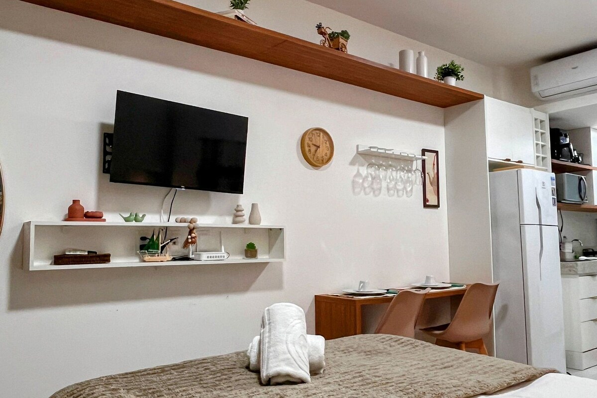 A hotel room with a bed, a TV, a desk with a chair, a clock, and a shelf with decorative items.