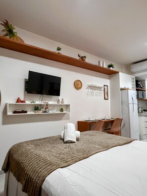 A hotel room with a bed, a TV, a desk with a chair, a clock, and a shelf with decorative items. - Mahrak Villa - VSH (Ipojuca)