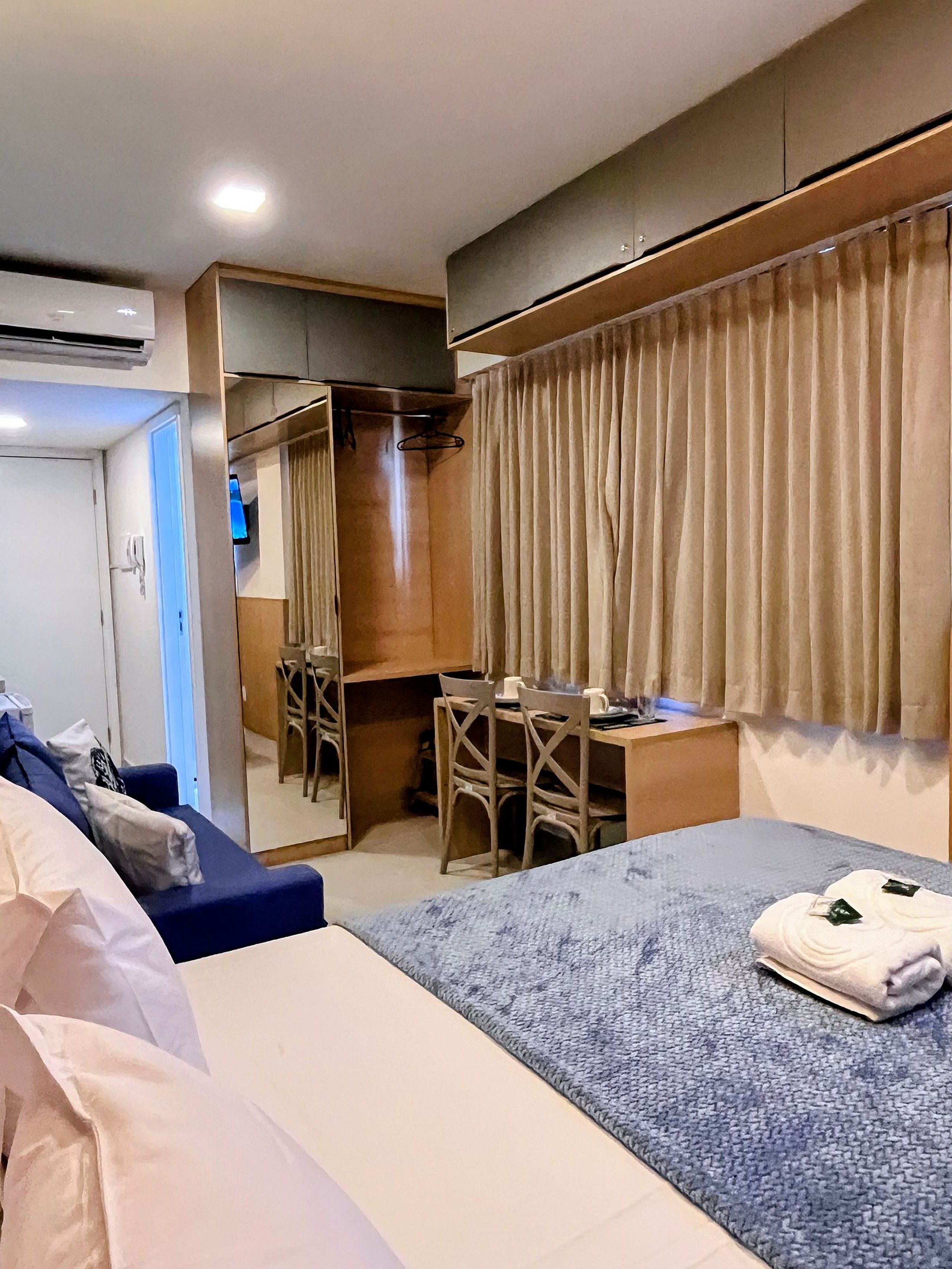 A hotel room with a bed, a desk, a chair, a wardrobe, and an air conditioning unit.