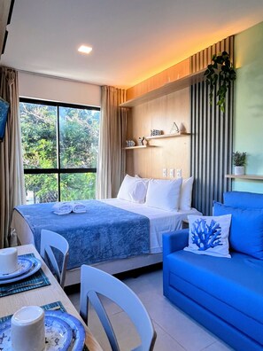 Una habitación de hotel con cama, un sofá azul, una mesa de comedor con platos y una taza, y una ventana con cortinas.