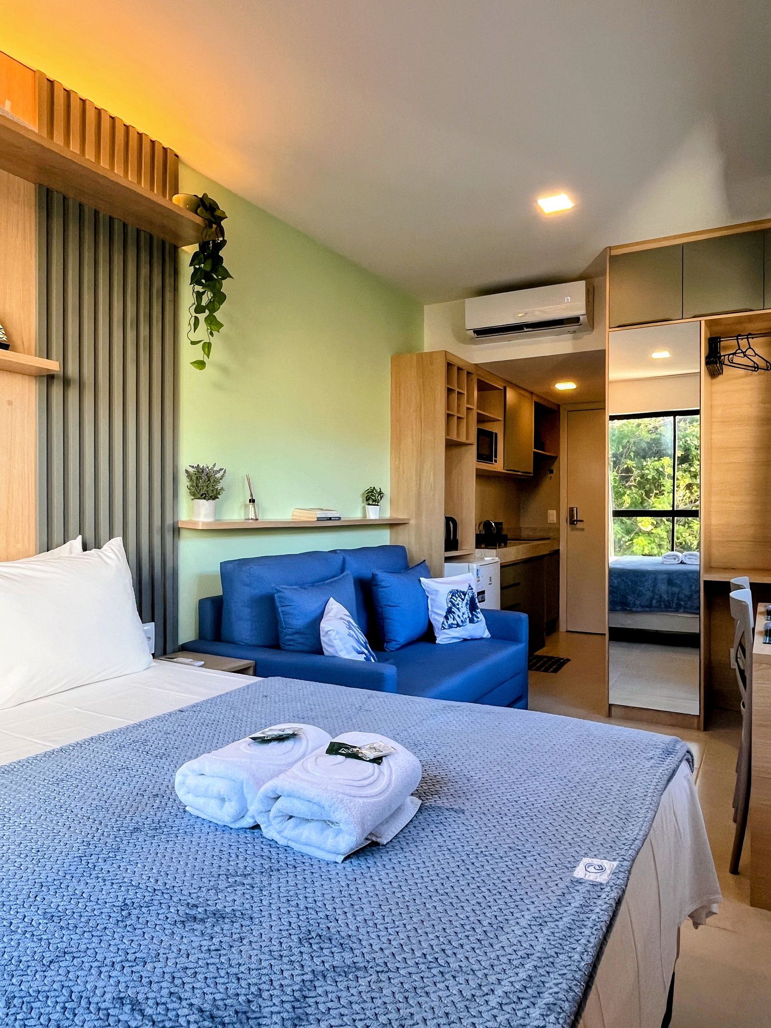 Une chambre d’hôtel moderne avec un lit, un canapé bleu, une petite kitchenette et une vue sur l’extérieur.