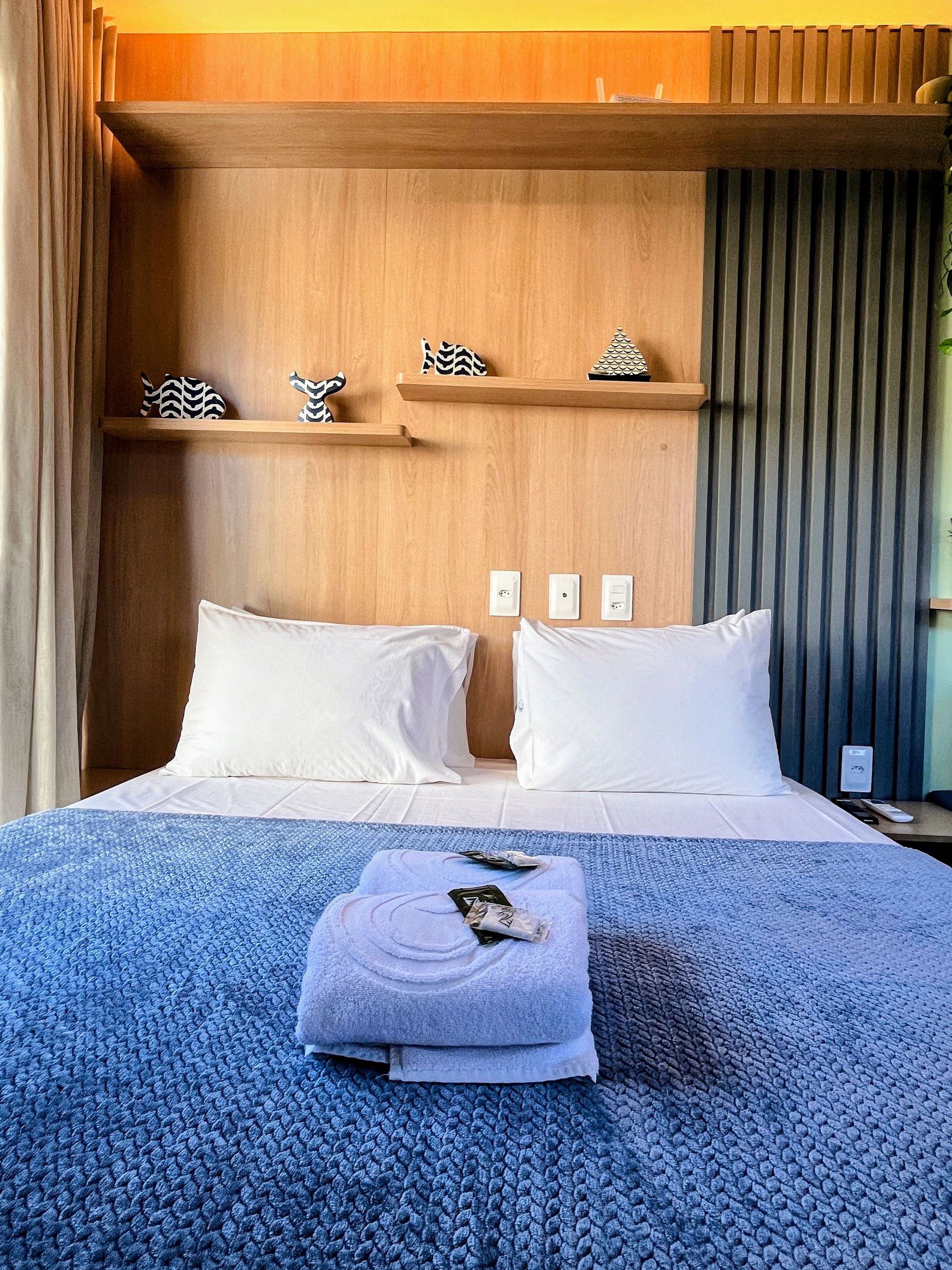 Een hotelkamer met een bed, kussens, een blauwe gebreide deken, een opgevouwen handdoek en een houten hoofdbord met planken.