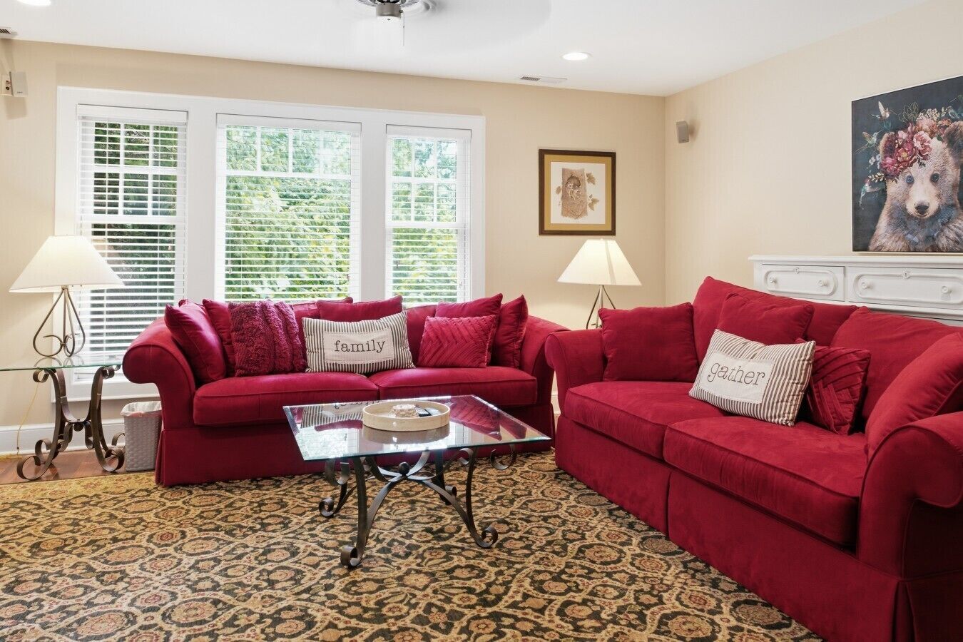 A living room with two red sofas, a glass coffee table, patterned carpet, two lamps, and a painting on the wall.