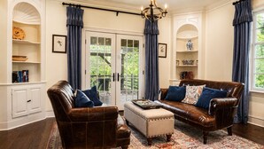 A living room with a brown leather sofa, a matching armchair, a small ottoman, and a chandelier. There are two windows with blue curtains, a white cabinet, and a bookshelf. - Hawk’s Landing | Hot Tub, Views & Game Room (Banner Elk)