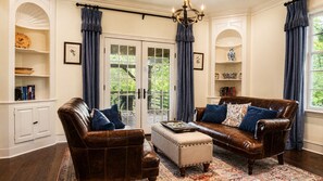 Ein Wohnzimmer mit einem braunen Ledersofa, einem passenden Sessel, einem kleinen Hocker und einer Kristallleuchtre. Zwei Fenster mit blauen Vorhängen, ein weißes Schränkchen und ein Bücherregal sind ebenfalls zu sehen.