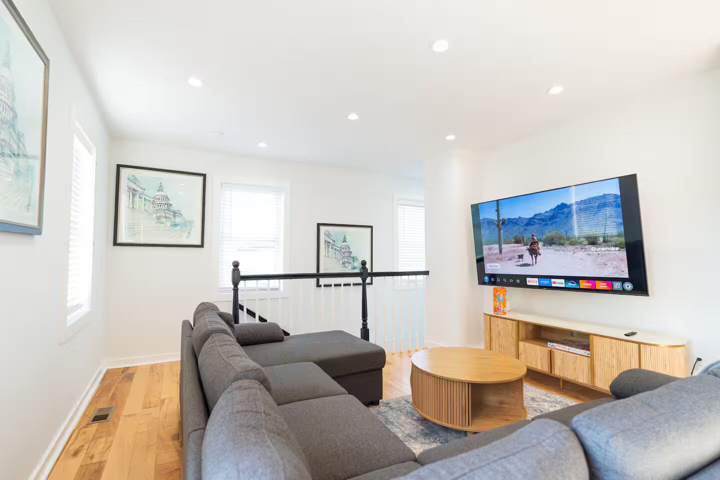 A modern living room with a large flat-screen TV, a grey sectional sofa, a wooden coffee table, and framed artwork on the walls.