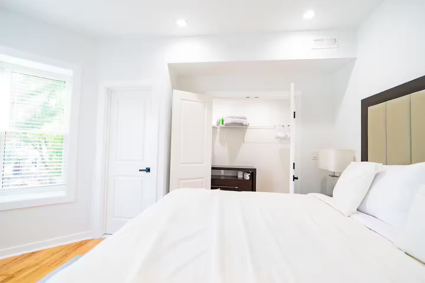 Una habitación de hotel con una cama grande, una mesita de noche, una lámpara y un ventanal con persianas.
