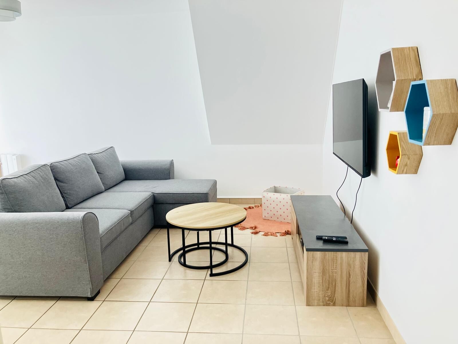 A modern living room with a grey sectional sofa, a round wooden coffee table, a TV on a wooden stand, and a geometric wall decoration.