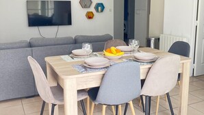 Ein Essbereich mit einem Holztisch, gedeckt für vier Personen, umgeben von Stühlen. Ein graues Sofa steht an der Wand, darüber ist ein Flachbildfernseher angebracht.