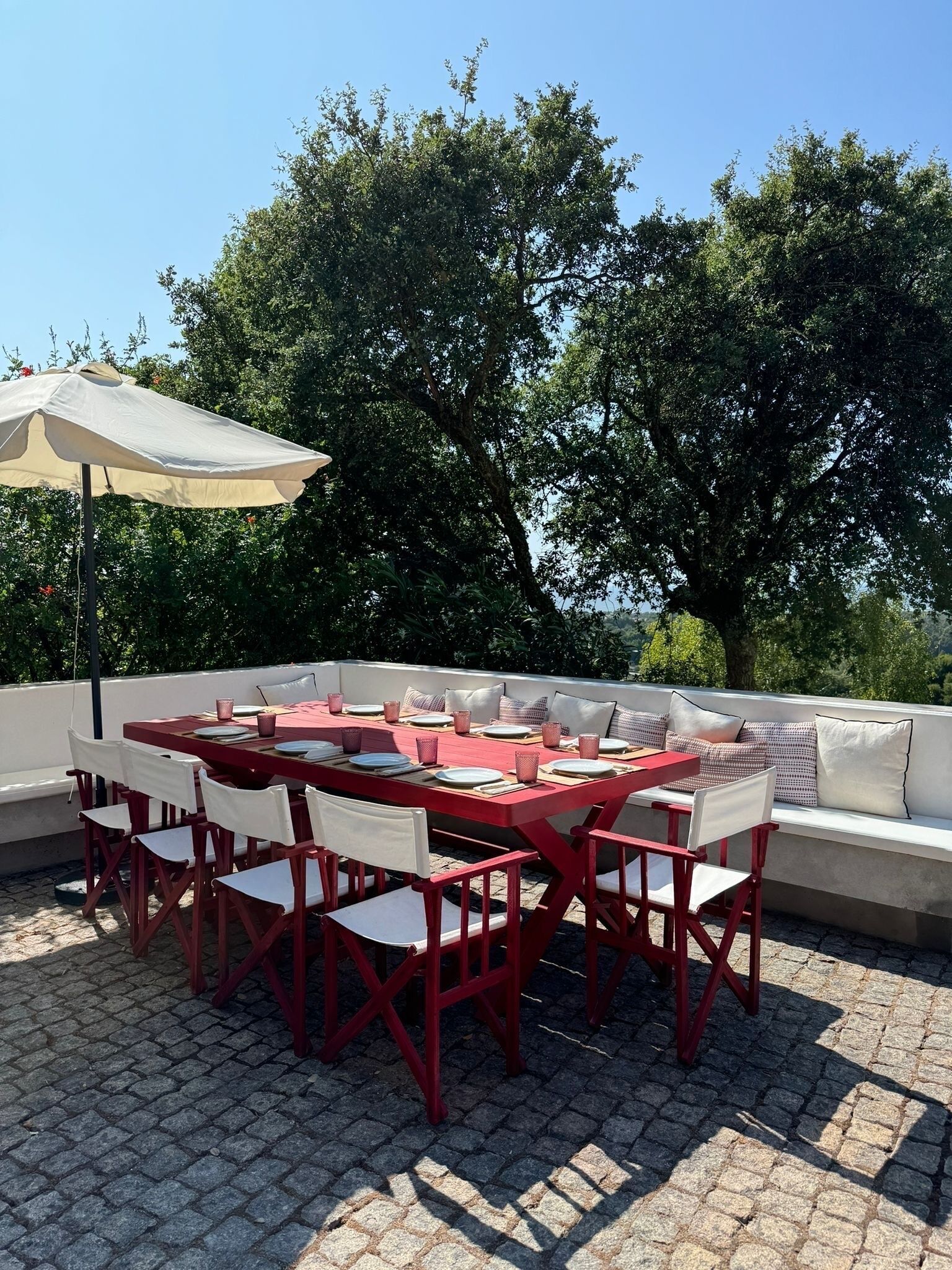 An outdoor dining area with a red table and white chairs, a beige umbrella, and a white cushioned bench.
