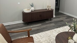 A modern living room with a flat-screen TV mounted on the wall, a dark wood console table with decorative items, a brown leather armchair, and a round coffee table with a white rug.