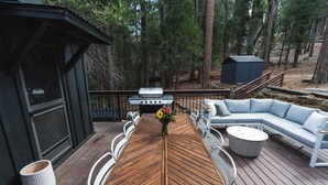 Eine Holzterrasse mit Esstisch, weißen Stühlen und einem grauen runden Tisch. Außerdem ist eine graue Outdoor-Sitzgruppe zu sehen.
