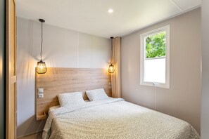 A bedroom with a wooden headboard, two bedside lamps, a window with a view of greenery, and a bed with patterned bedding.