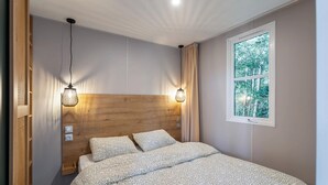 Une chambre à coucher avec une tête de lit en bois, deux lampes de chevet, une fenêtre donnant sur la forêt et un lit recouvert d’une literie à motifs.