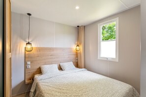 A bedroom with a wooden headboard, two bedside lamps, a window with a view of greenery, and a bed with patterned bedding.