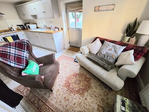 A living room with a sofa, a chair, and a patterned rug. There is a kitchen area with cabinets and a microwave. - Cosy tucked-away Barn conversion in Matlock Green (Matlock)