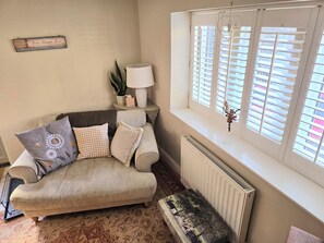 Ein gemütliches Wohnzimmer mit einer Couch, einem Beistelltisch mit Lampe, einem Blumentopf und einem Fenster mit Jalousien.