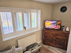Ein Wohnzimmer mit Fernseher, Holzschrank, Heizkörper und einem Fenster mit Jalousien.