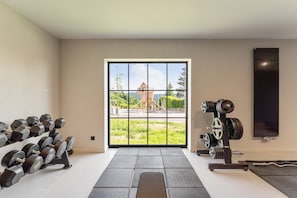 Ein Fitnessraum mit Trainingsgeräten, einem großen Fenster mit Blick auf ein Gebäude und Grünflächen, sowie einer Tür.