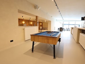 A pool table in a room with a kitchen area and a view to the outside.