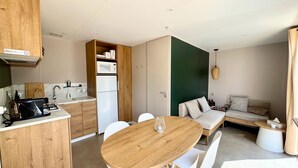 Una cocina moderna con armarios de madera, microondas integrado y refrigerador blanco. Hay un comedor con mesa de madera y sillas blancas.