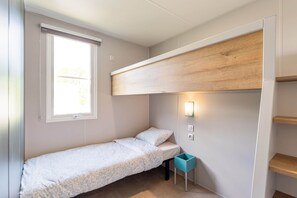 A small, single-bed room with a window, a wall-mounted light, and a wooden shelf.