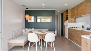 A modern kitchen with wooden cabinets, a dining area with a round table and white chairs, and a built-in bench with cushions. - Camping Papillon Bouillon - Premium Quiet (Bouillon)