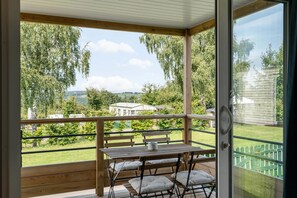 Een houten terras met een tafel en stoelen, met uitzicht op een groen landschap met bomen en een gebouw in de verte.