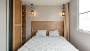A bedroom with a wooden headboard, two bedside lamps, a bed with patterned bedding, and a large window with curtains. - Camping Papillon Bouillon - Premium Quiet (Bouillon)