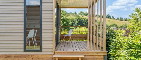 Une terrasse en bois avec un banc et des chaises, une vue sur un paysage verdoyant et une fenêtre avec une chaise à l’intérieur.