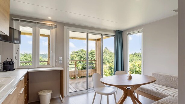Een moderne keuken met een ingebouwde televisie, een eethoek met een ronde tafel en stoelen, en een balkon met een houten terras en uitzicht op groen.