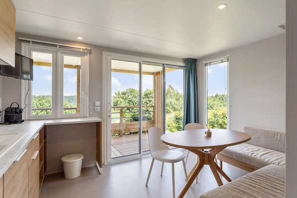 Eine moderne Küche mit integriertem Fernseher, ein Essbereich mit rundem Tisch und Stühlen sowie ein Balkon mit Holzdeck und Blick ins Grüne.