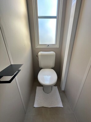 A small, narrow bathroom with a toilet, a window, and a black shelf. - Camping Papillon Kinrooi - 88 (Kinrooi)