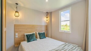 A bedroom with a wooden headboard, two bedside lamps, a large window with a view, and a bed with patterned bedding. - Camping Papillon Kinrooi - La Casetta (Kinrooi)