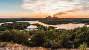 Panorama di un lago al tramonto, circondato da una fitta foresta e un piccolo edificio.