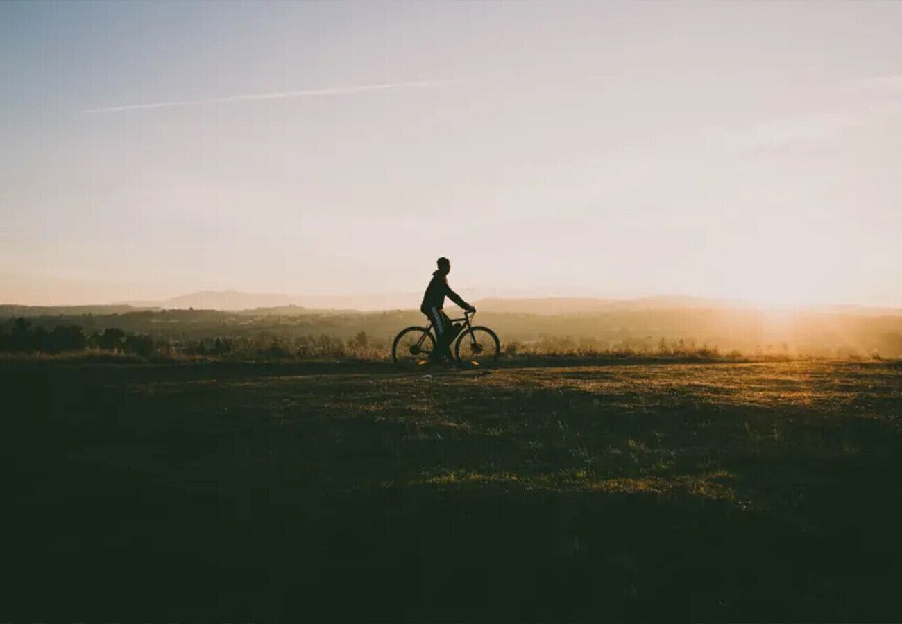 Silhouet van een persoon die op een fiets rijdt over een vlak, open veld tijdens de zonsondergang.