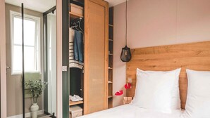 Una camera da letto con una testiera in legno, un letto con biancheria bianca, un armadio pieno di vestiti e una pianta in vaso vicino alla finestra.