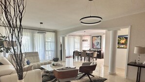 A spacious living room with a white sofa, a coffee table, and a dining area with chairs and a table. - PGA Golf Course 3-bedroom condo AC & VIEWS- steps to the Beach, Clubhouse & Pool (Río Grande)
