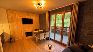 Een gezellige kamer met een houten wand, een flatscreen televisie, een eettafel met stoelen, een klein rond tafeltje met planten en een bank.